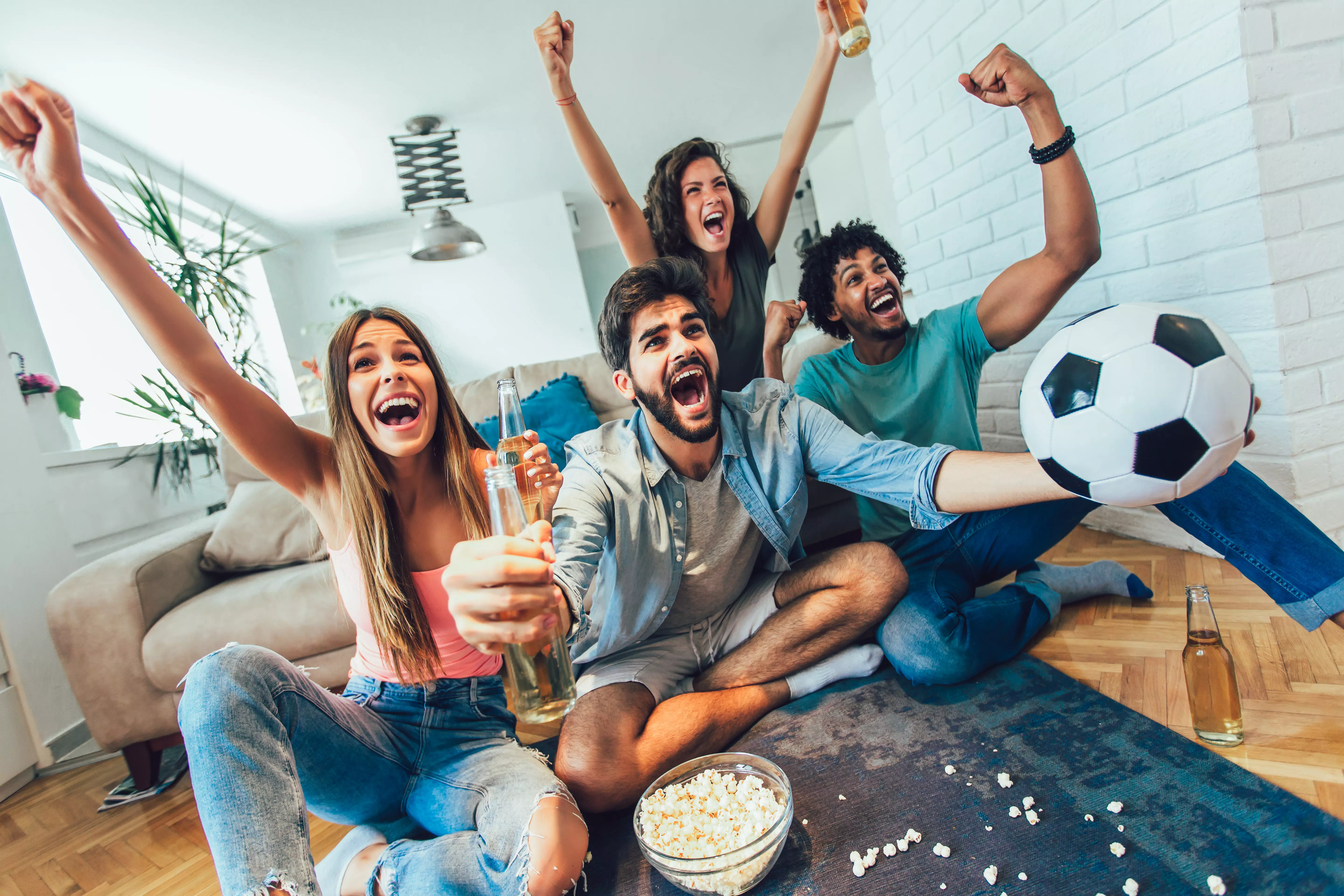 Jubelnde Fußballfans im Wohnzimmer, mit einem Fußball und Popcorn auf dem Tisch, eingefangen in einem Moment der Freude und des gemeinschaftlichen Miteinanders, perfekt für Fußballliebhaber, präsentiert von flaggenmeer