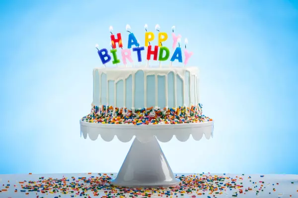 Farbenfrohe Geburtstagstorte mit 'Happy Birthday' als Kerzen, auf einem hellblauen Hintergrund mit mittig hellerer Ausleuchtung, dekoriert mit Smarties am unteren Rand der Torte und auf dem Tisch, eine fröhliche Darstellung für Geburtstagsfeiern, angebote